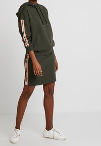 Olijfgroene hoodie met roze strepen op de mouwen, gecombineerd met een bijpassende rok. Witte sneakers maken de outfit compleet. Zachte stofstructuur.