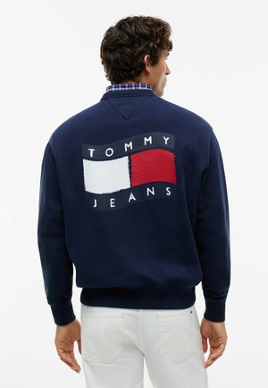 Mężczyzna w granatowej bluzie z "Tommy Jeans" i czerwono-białym logo flagi na plecach, w połączeniu z białymi spodniami.