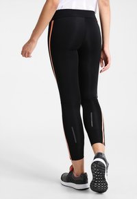 Černé legíny z pružného materiálu, s oranžovými bočními pruhy, reflexními akcenty a tapered střihem. Nosené s atletickými botami.