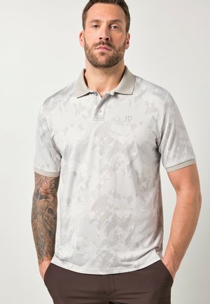 Mand med skæg og tatoveret venstre arm iført lysegrå polo med mønster og mørke bukser, stående med hænderne i lommerne.