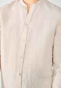 Beige linnen shirt met een mandarijnkraag, voorzien van een volledige knoopsluiting en subtiele textuur. Knopen zijn licht van kleur en rond.