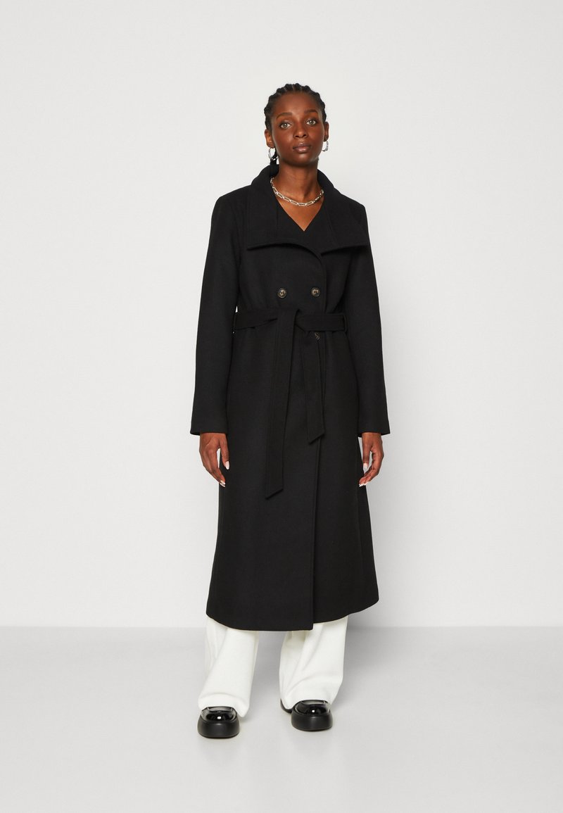 Cappotto lungo nero con design doppiopetto, colletto ampio, cintura in vita e bottoni prominenti. Indossato sopra pantaloni bianchi a gamba larga.