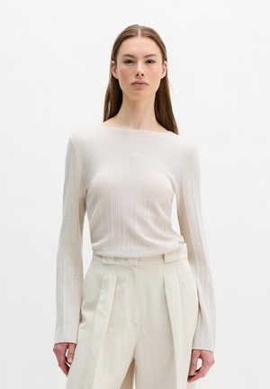 Vrouw met steil bruin haar, gekleed in een wit geribd shirt met lange mouwen en een hooggetailleerde crème plissébroek, staande tegen een effen achtergrond.