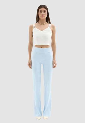 Donna che indossa un top corto bianco con doppie bretelle, pantaloni larghi a vita alta color azzurro chiaro, in piedi con le braccia lungo i fianchi.