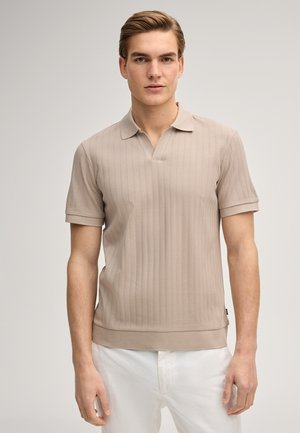 Jonge man met lichtbruin haar, gekleed in een beige korte mouw gebreide poloshirt en witte broek, tegen een effen lichte achtergrond.