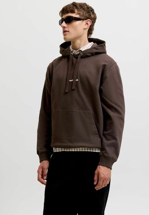 Jeune homme portant un sweat à capuche marron foncé par-dessus une chemise à carreaux et un pantalon noir, avec les cheveux courts et des lunettes de soleil foncées, debout devant un fond uni.