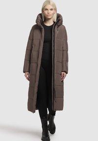 Rjav podložen dolg plašč s kapuco, ki ima zadrgo spredaj, stranske žepke in quiltan design. Nosi se čez črn zgornji del in hlače.