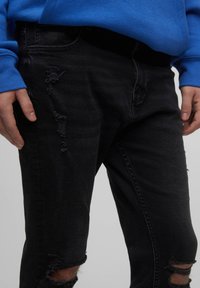 Ruce spočívají u boků, oblečeny v černých roztrhaných džínach a modrém topu s dlouhým rukávem, na jednobarevném pozadí.