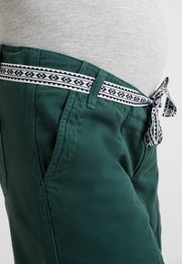 Green maternity pants featuring a decorative black and white patterned waistband and tie. Smooth fabric with side pockets.