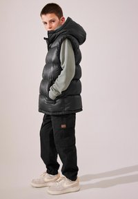 Chaleco de plumas negro con capucha, camiseta de manga larga texturizada gris debajo, pantalones cargo negros y zapatillas de color claro.