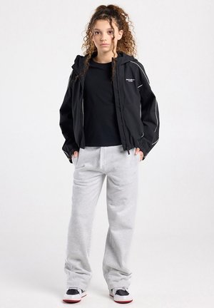 Jongere met krullend haar die een zwart gewatteerd jack, zwart T-shirt, grijze joggingbroek en zwart-witte sneakers draagt, staand met handen in de zakken.