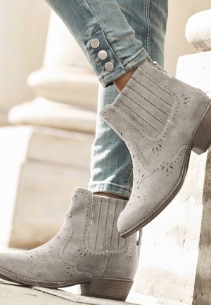 Graue Wildleder-Ankle Boots mit spitzem Zehenbereich, detaillierten Ausschnitten, vertikalen Nähten und einem niedrigen Holzabsatz, kombiniert mit hellen Jeans.