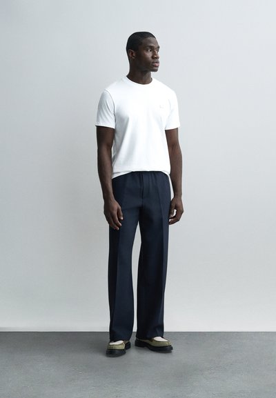 Άνδρας που φοράει λευκό κοντομάνικο μπλουζάκι, ναυτικό μπλε παντελόνι φαρδύ στη μέση, και μπεζ με μαύρα παπούτσια, στέκεται μπροστά σε ανοιχτό γκρι φόντο.