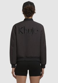 Must must jacket, mille "Khujo" logo tikandiga seljal, soonikust mansetid ja alläär, sile tekstuur ja lühike disain.