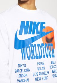 Valkoinen T-paita, jossa on ylisuuri sininen "NIKE"-logo ja "WORLDTOUR"-teksti, sekä mustalla listatut kaupunkien nimet alla. Sujuva kankaan rakenne.