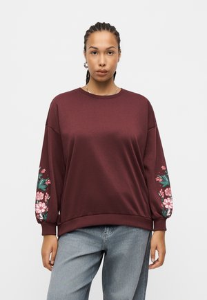 Kvinde vendt fremad i en bordeaux sweatshirt med blomster-mønstre på ærmerne, lyseblå jeans, håret i fletninger, neutral baggrund.