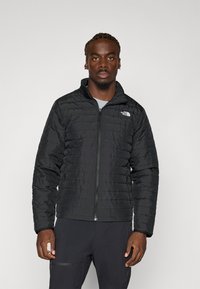 Juodas pūkinis švarkas su užtrauktuvu priekyje, pasižymintis aukštu kaklu ir tekstūruota paviršiumi. "The North Face" logotipas matomas kairiajame krūtinės šone.