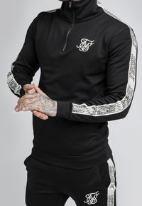 Muž v černém sportovním obleku s bílými vzorovanými pruhy na rukávech a nohavicích, s rukama sepjatýma, ukazujícími tetování na prstech.