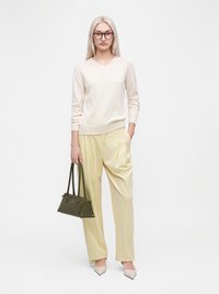 Femme aux longs cheveux blonds et lunettes porte un pull crème, un pantalon jaune pâle, des chaussures beiges, tenant un sac à main vert foncé d'une main.