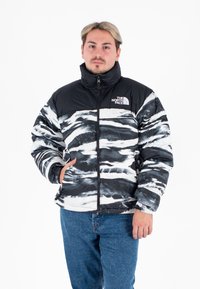 Chaqueta acolchada en negro y blanco con patrón abstracto de olas, cierre de cremallera, cuello alto y logotipo en el lado izquierdo del pecho. Textura suave y acolchada.