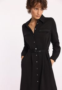 Czarna sukienka koszulowa z długimi rękawami, dwoma kieszeniami na piersi, zapięciem na guziki i materiałowym paskiem. Posiada teksturowany materiał oraz metalowe guziki.