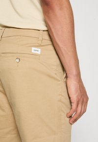 Des pantalons en coton beige présentent une poche arrière avec un bouton, une étiquette de logo embossée et une coupe slim. Le tissu a une texture lisse.