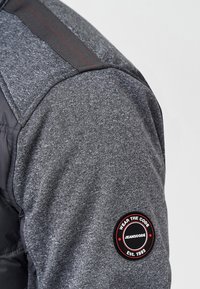 Grauer, strukturierter Jackenärmel mit schwarzem und rotem rundem Aufnäher, auf dem am Oberarm "Wear the Code Jeanscode Est. 1993" steht.