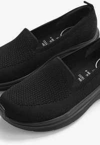 Schwarze Slip-on-Strickschuhe mit strukturierter Oberfläche, gepolsterter Sohle und Zuglasche an der Ferse für einfaches Anziehen.
