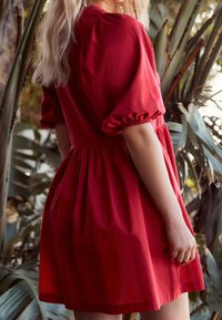 Robe en coton rouge avec manches bouffantes, corsage ajusté et jupe froncée. Texture lisse, longueur au genou, sur fond de verdure.