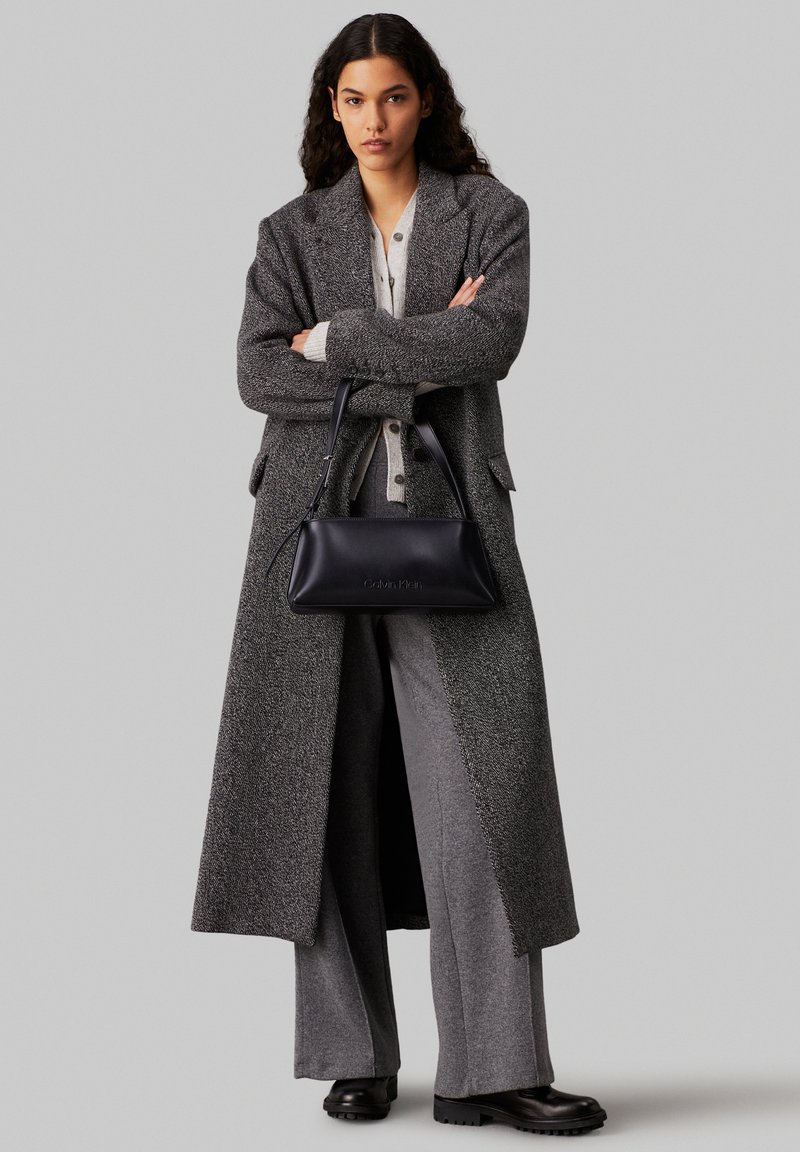 Cappotto lungo grigio texturizzato su un maglione grigio chiaro, abbinato a pantaloni larghi grigi e una borsa in pelle nera con un branding discreto.