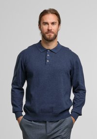 Mann mit Bart und zurückgebundenem Haar, der einen marineblauen Langarm-Polo-Pullover und graue Hosen trägt und mit den Händen in den Taschen steht.