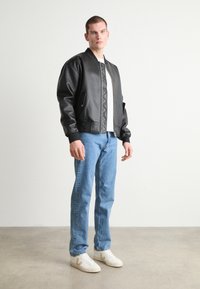 Jachetă bomber din piele ecologică neagră, cu manșete și guler striate, având față cu fermoar și detalii quilted. Asortată cu blugi denim de culoare albastră deschisă și adidași albi.