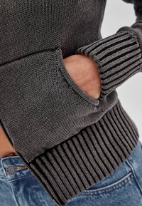 Szary sweter o fakturowanym splocie, z przednią kieszenią i prążkowanymi mankietami. Noszony z dżinsami, na których widoczny jest pas.