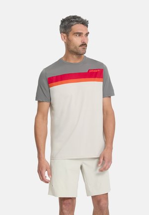 Homme portant un T-shirt à manches courtes avec blocs de couleurs gris, rouge et orange et un short beige clair, debout et regardant sur le côté.