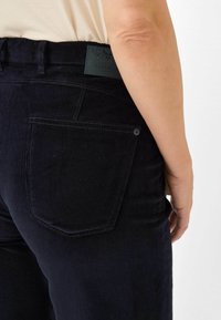 Donkerblauwe jeans met een gestructureerd oppervlak, voorzien van een achterzak en een leren label met de tekst "RAPHAELA." Subtiele stikdetails zijn zichtbaar.