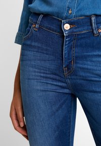 Jeans ajustados de denim azul con botones de remache de cobre, costuras de color naranja claro y un diseño de bolsillo frontal, mostrados desde la cintura hasta la mitad del muslo.