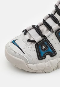 Zapatilla blanca con acentos en negro y azul, que presenta una suela de goma texturizada, paneles de malla y un prominente logo de 'A'.
