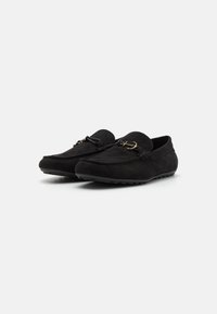 Mocassins en suede noir avec une texture lisse, présentant un bout rond, des coutures décoratives et un accent en métal doré sur le devant.