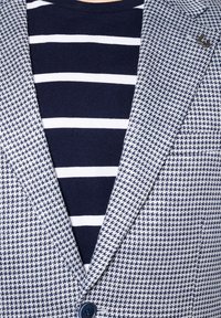 Granatowo-biała w paski koszulka pod marynarką w desenie houndstooth w odcieniach niebieskiego. Marynarka ma detal z guzikami i klapami.