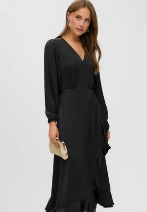 Femme aux longs cheveux bruns portant une robe noire à manches longues et décolleté en V, tenant une petite pochette dorée sur un fond uni.