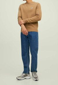 Ruskea neulepaita, jossa on pyöreä kaula-aukko, yhdistettynä sinisiin suoriin farkkuihin ja harmaisiin lenkkareihin, joissa on valkoiset korostukset ja nauhat.