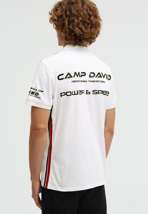 Mann mit lockigem Haar, der ein weißes Poloshirt mit schwarzen und roten Streifen an den Seiten trägt; auf der Rückseite steht der Text "CAMP DAVID POWER & SPEED".