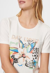 Camiseta de manga corta en color crema con un diseño floral colorido con acentos en azul y negro, texto "DAY LIGHT" adornado y una textura suave.