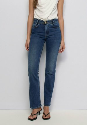 Mujer con vaqueros azules ajustados con un pequeño cinturón, top blanco sin mangas y sandalias negras de tacón, de pie frente a un fondo liso.