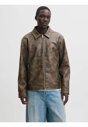 Jeune homme portant une veste en cuir brun marbrée zippée, un jean bleu clair et de petites boucles d'oreilles dorées en forme d'anneau, debout devant un fond uni.
