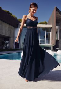 Robe maxi bleu marine avec corsage en dentelle et bretelles spaghetti, tissu fluide, portée avec une pochette, près d'une piscine et d'une architecture moderne.