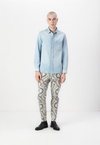 Svijetloplena traper košulja s dugim rukavima i džepom na prsima, uparena s krem hlačama koje imaju crne paisley uzorke. Crne gležnjače dopunjuju ovaj outfit.