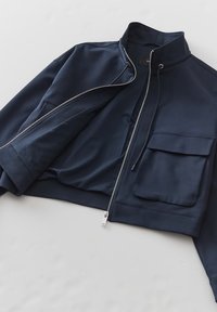 Marinblå kort jacka i slät tyg, med hög krage, dragkedja fram och en bröstficka på vänster sida.