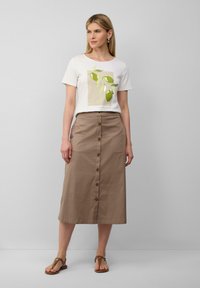 Witte t-shirt met groene olijfafbeelding, gecombineerd met een knielange bruine knopenrok. Platte bruine sandalen maken de outfit compleet.