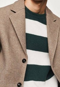 Beżowa marynarka jodełkowa z klapowanym kołnierzem i czarnymi guzikami, noszona na zielono-białym swetrze w paski.